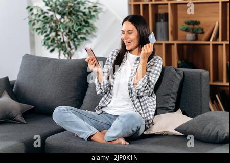 Une femme brune caucasienne satisfaite fait des achats sur Internet en choisissant des marchandises dans des magasins en ligne, s'assoit sur un canapé dans le salon, tient un smartphone et une carte bancaire, paie pour les marchandises, sourit Banque D'Images