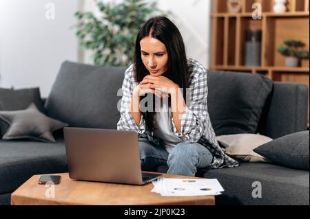 Focalisé confiance caucasien brune femme, s'asseoir dans le salon, à l'aide d'un ordinateur portable, travaillant sur un projet important de la maison, concentré regarde l'écran, à la recherche d'informations sur l'Internet Banque D'Images