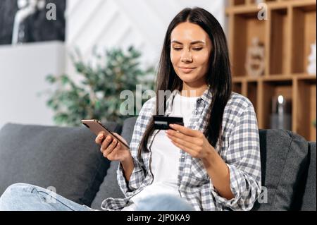 Achats en ligne. Une femme caucasienne satisfaite fait des achats sur Internet en choisissant des marchandises dans des magasins en ligne, s'assoit sur un canapé dans le salon, tient un smartphone et une carte bancaire, paie pour les marchandises, sourire Banque D'Images