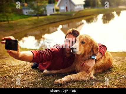 Il aime prendre des photos. Prise de vue en longueur d'un beau jeune homme et de son chien prenant des selfies au bord d'un lac dans le parc. Banque D'Images