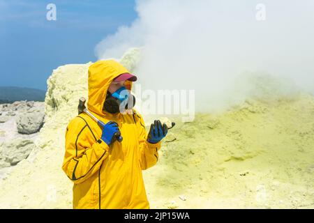 une femme volcanologue, à l'arrière-plan d'un fumarale fumeur, examine un échantillon de minerai de soufre Banque D'Images