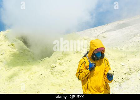une scientifique volcanique femelle, sur le fond d'un fumarale fumeur, examine un échantillon d'un minerai de soufre Banque D'Images