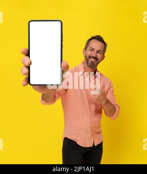 Joyeux homme mi-âge gris à cheveux tenue tenant un grand smartphone pointant le doigt dessus portant une chemise pêche isolée sur jaune. Homme mûr musclé avec publicité d'application de téléphone. Maquette d'écran blanc. Banque D'Images