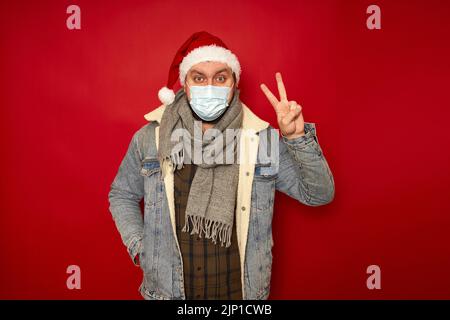 Homme en chapeau de Noël, masque médical sur le visage montre la paix signe de victoire main isolé arrière-plan de studio Banque D'Images