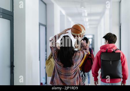 Vue arrière des jeunes élèves du secondaire qui marchent dans le couloir à l'école avec ballon de basket-ball, concept de retour à l'école. Banque D'Images