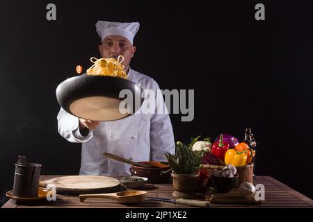 Chef masculin en uniforme blanc tenant une poêle à frire, sautant des spaghetti avec des légumes frais volant dans l'air avant de servir dans un restaurant Banque D'Images