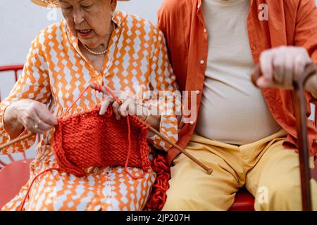 Femme âgée assise dans le jardin sur le banc avec son mari et tricoter le foulard rouge. Banque D'Images
