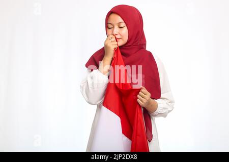 Jeune femme asiatique musulmane embrassant le drapeau rouge et blanc dans des vêtements rouges et blancs célébrant le jour de l'indépendance indonésienne isolée sur fond blanc Banque D'Images