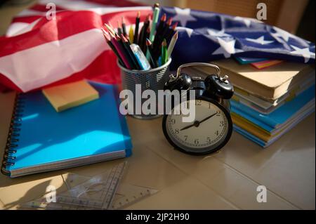 Réveil noir, fournitures scolaires, papeterie et manuels empilés sur une table avec drapeau américain sur le fond. Banque D'Images