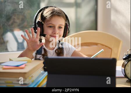 Une jeune fille intelligente de l'école primaire portant un casque sans fil regardant une leçon en ligne sur une tablette numérique. Enseignement à domicile Banque D'Images