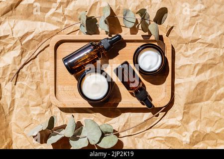 Bouteilles cosmétiques en verre foncé avec compte-gouttes et crème faciale sur fond de papier artisanal avec feuilles d'eucalyptus. Concept de cosmétiques naturels, Essen naturel Banque D'Images