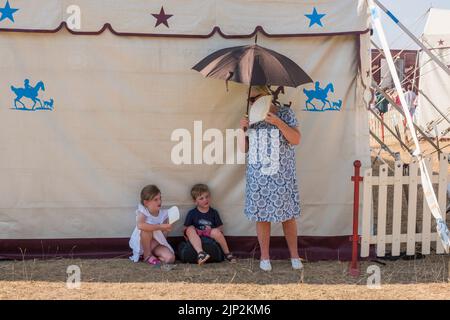 Une famille trouve une place à l'ombre et essaie de se rafraîchir en utilisant des ventilateurs de papier et un parapluie, tout en attendant dans la chaleur brûlante pour voir le Cirque de Gifford sur Minchinhampton Common, Gloucestershire, Royaume-Uni, pendant la vague de chaleur d'août 2022 Banque D'Images