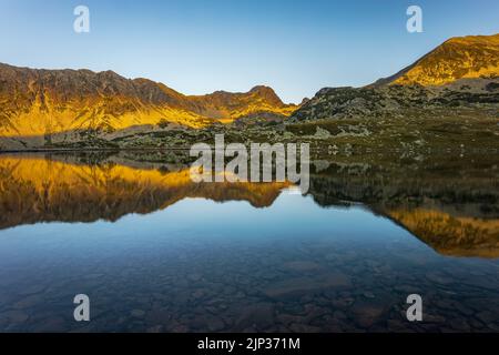 Lever de soleil avec lumière douce et orange illuminant les sommets des montagnes de Retezat et se reflétant dans le lac Bucura. Photo prise le 7th août 2022 sur la rive Banque D'Images