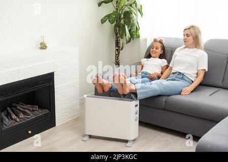 Chauffage électrique à la maison. Une jeune famille réchauffe ses mains gelées près d'un radiateur de chauffage Banque D'Images