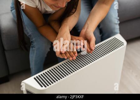 Chauffage électrique à la maison. Une jeune famille réchauffe ses mains gelées près d'un radiateur de chauffage Banque D'Images