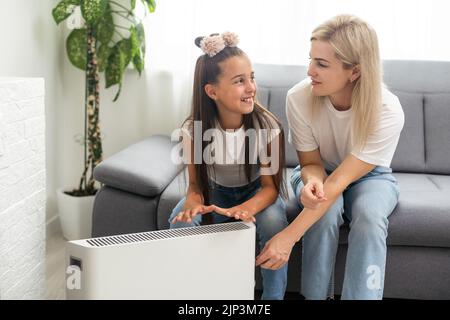 Chauffage électrique à la maison. Une jeune famille réchauffe ses mains gelées près d'un radiateur de chauffage Banque D'Images