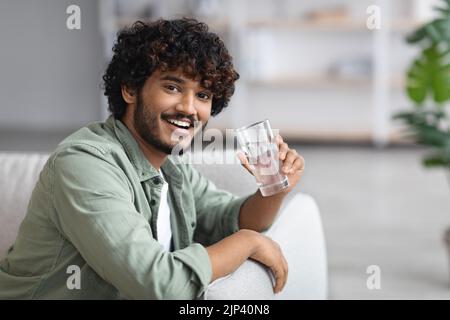 Homme à la peau sombre qui boit de l'eau et souriant, intérieur de la maison Banque D'Images