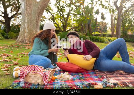 Heureux couple biracial ayant pique-nique assis sur un tapis et boire du vin dans le jardin d'automne et souriant Banque D'Images