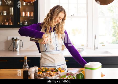 Bonne femme caucasienne debout dans la cuisine en tablier mettant de l'huile sur des légumes hachés Banque D'Images