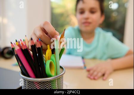 Concentrez-vous sur un support avec des crayons colorés, des ciseaux, de la papeterie sur la table contre un garçon d'école flou en train de sortir un stylo Banque D'Images