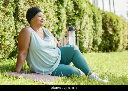 Vue latérale sur toute la longueur d'une femme noire en surpoids se détendant sur l'herbe tout en s'faisant de l'exercice à l'extérieur avec une bouteille d'eau, un espace de copie Banque D'Images