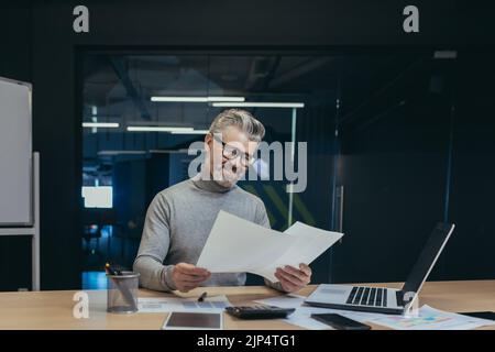 Homme d'affaires mûr et souriant derrière un travail papier, patron à poil gris travaillant avec des comptes et des documents, homme travaillant dans un bureau moderne, investisseur prospère examinant les rapports financiers Banque D'Images