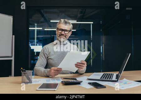 Portrait d'un financier chevronné, d'un homme travaillant avec des documents à l'intérieur d'un immeuble de bureaux, d'un homme d'affaires souriant et regardant un appareil photo tenant des documents et des factures, d'un patron travaillant à l'aide d'un ordinateur portable Banque D'Images
