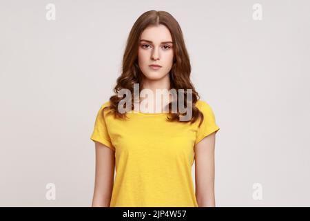 Portrait d'une femme de jeune âge à poils bruns assez sérieuse et bossy, en T-shirt jaune décontracté, regardant l'appareil photo avec suspicion et accusant. Prise de vue en studio isolée sur fond gris. Banque D'Images