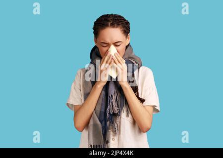 Portrait d'une femme malade avec des dreadlocks noirs éternuements dans un mouchoir, debout enveloppé dans un foulard, attrapant le froid, symptômes de la grippe, portant une chemise blanche. Studio d'intérieur isolé sur fond bleu. Banque D'Images
