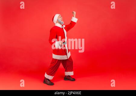 Portrait complet d'homme âgé avec barbe grise portant le costume du père noël criant pour la joie avec les poings relevés, célébrant les vacances, champion. Studio d'intérieur isolé sur fond rouge. Banque D'Images