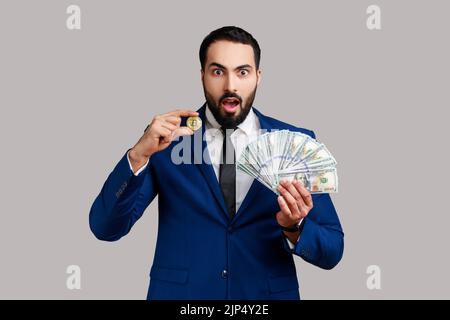 Un homme choqué montrant des billets en dollars et une pièce de monnaie en or de crypto-monnaie, transférant de l'argent dans des bitcoins numériques, portant un costume de style officiel. Prise de vue en studio isolée sur fond gris. Banque D'Images