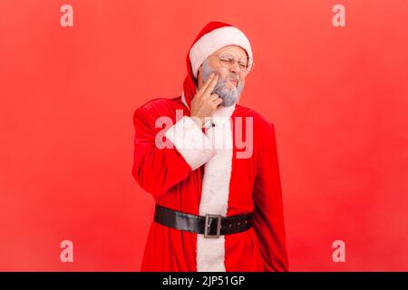 Portrait d'un homme malade âgé avec une barbe grise portant le costume du père noël debout et touchant sa joue, souffrant d'une terrible douleur à la dent. Studio d'intérieur isolé sur fond rouge. Banque D'Images