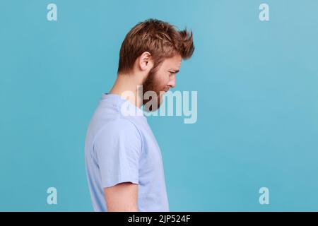 Vue latérale du jeune homme en colère barbu debout et regardant la caméra avec le visage de tristesse insatisfait, exprimant la douleur, ayant mauvaise humeur. Studio d'intérieur isolé sur fond bleu. Banque D'Images