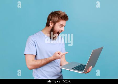 Portrait d'un beau homme surpris surpris barbu pointant le doigt sur l'écran d'un ordinateur portable avec de grands yeux choqués par le contenu. Studio d'intérieur isolé sur fond bleu. Banque D'Images