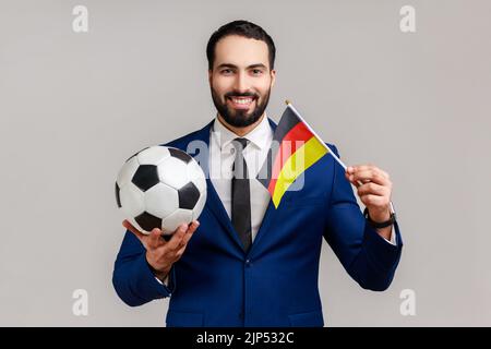 Homme barbu positif tenant le drapeau de l'allemagne et de football noir et blanc classique ballon et match de surveillance, portant le style officiel de costume. Prise de vue en studio isolée sur fond gris. Banque D'Images