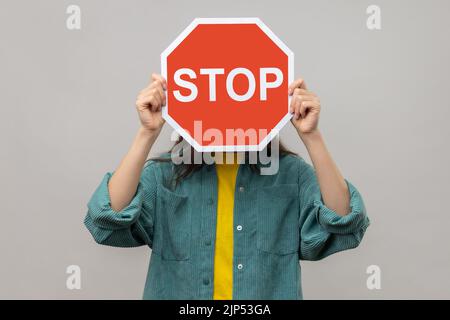 Attention, manière interdite. Personne inconnue se cachant face derrière le symbole Stop, signalisation routière rouge signalant un accès restreint, service interdit, porter une veste de style décontracté. Prise de vue en studio isolée sur fond gris. Banque D'Images