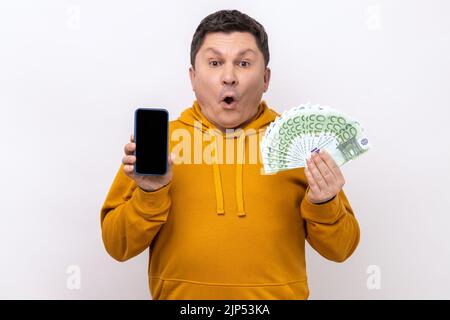 Surpris homme tenant un smartphone à écran vide, un fan de publicité de cent euros, des opérations bancaires et des gains en ligne, portant un sweat à capuche de style urbain. Studio d'intérieur isolé sur fond blanc. Banque D'Images