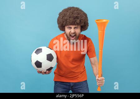 Portrait d'un homme avec une coiffure afro portant un T-shirt orange debout avec une balle de football et une corne, criant joyeusement avec une expression folle. Studio d'intérieur isolé sur fond bleu. Banque D'Images