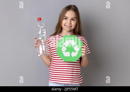 Portrait d'une petite fille souriante satisfaite portant un T-shirt rayé tenant une bouteille de plastique vide et une affiche de recyclage, pensant vert. Prise de vue en studio isolée sur fond gris. Banque D'Images