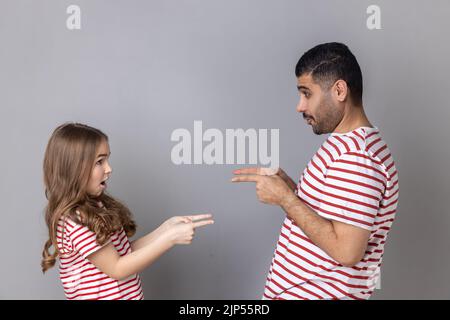 Portrait d'un père et d'une fille étonnés en T-shirts rayés se pointant les uns vers les autres, regardant avec les grands yeux, choisissant le tour. Prise de vue en studio isolée sur fond gris. Banque D'Images