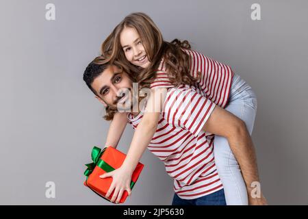 Portrait d'un père et d'une fille extrêmement heureux en T-shirts rayés célébrant ensemble les vacances, papa tenant un enfant avec une boîte cadeau sur son dos. Prise de vue en studio isolée sur fond gris. Banque D'Images