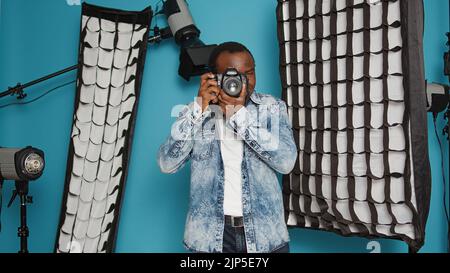 Photographe de sexe masculin utilisant un appareil photo reflex numérique et un objectif pour prendre des photos dans les coulisses d'un studio, en utilisant un équipement professionnel. Utilisation de la vue mise au point pour photographier des images sur l'arrière-plan. Banque D'Images