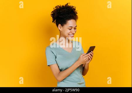 Jeune femme afro-américaine heureuse et heureuse avec des cheveux bouclés, en utilisant son smartphone, en regardant l'écran, en surfant sur Internet, en discutant avec des amis, debout sur un arrière-plan orange isolé, souriant Banque D'Images