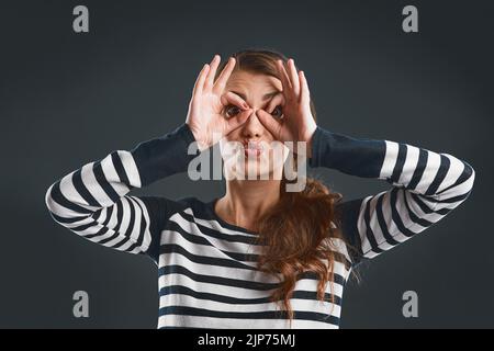 Elle égayera votre journée. Portrait studio d'une jeune femme gaie faisant un visage avec ses mains tout en se tenant contre un arrière-plan sombre. Banque D'Images