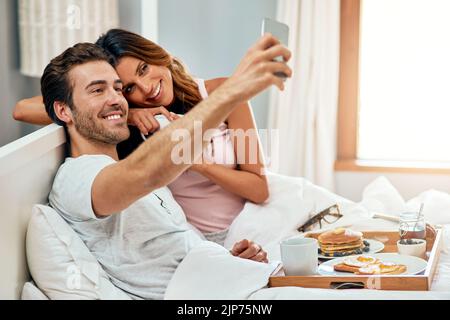 Bonjour. Un jeune couple heureux prenant un selfie tout en prenant le petit déjeuner au lit à la maison. Banque D'Images