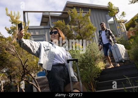 Divers couple prenant un selfie avec des bagages et souriant sur des escaliers en bois Banque D'Images