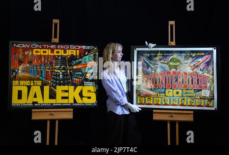 Photo non publiée du 02/08/22 d'un employé de Propstore qui ajuste une affiche autographiée de Sylvia Anderson, Gerry Anderson et David Lane du film 1960 « Thunderbirds are Go » (estimation £600 - 800) exposée en même temps qu'une affiche Quad du Royaume-Uni du film 1965 « Dr Who and the Daleks » (Estimation £1 500 - 2 500) lors d'un aperçu de leur prochaine vente aux enchères d'affiches à leur maison de vente aux enchères à Rickmansworth, Hertfordshire. Date de publication : mardi 16 août 2022. Banque D'Images