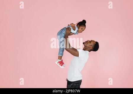 Bonne paternité. Joyeux papa africain tenant sa jolie fille sur les mains prétendant qu'elle vole, fille se propageant les mains, imitant avion, fond rose studio. Banque D'Images