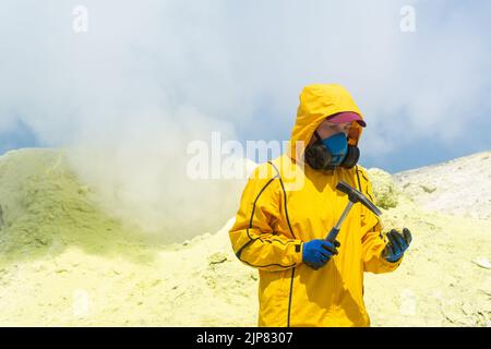une scientifique volcanique femelle, sur le fond d'un fumarale fumeur, examine un échantillon de minerai de soufre avec un marteau géologique Banque D'Images