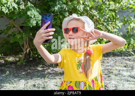 Portrait d'une petite fille qui prend un selfie par smartphone dans le parc d'été. Une petite fille gaie dans une robe jaune, chapeau blanc et lunettes de soleil prend un Banque D'Images